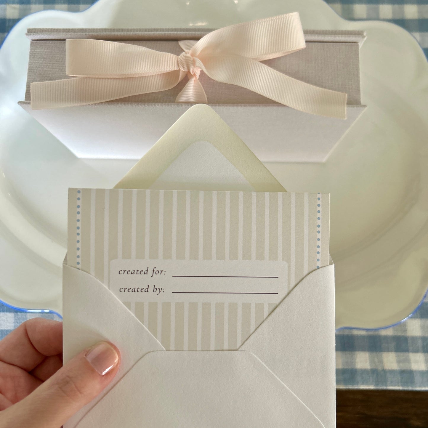White needlepoint card with a pink bow held by a hand on a checkered tablecloth.